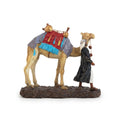 Figurine of a person in traditional attire leading a pack camel with a red saddl